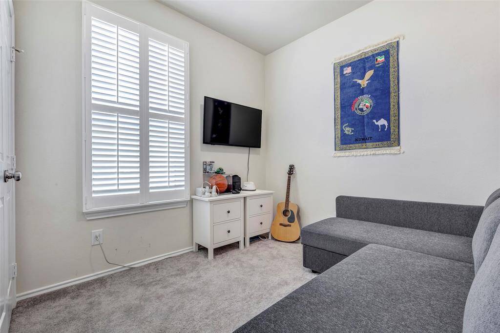 This is a cozy, well-lit room with a large window, plantation shutters, neutral wall color, and carpeted flooring. It includes a mounted TV, a small white cabinet, and a comfortable-looking gray sectional sofa. A decorative tapestry adds a touch of personality, and there's a guitar on the side, suggesting a creative or musical space.