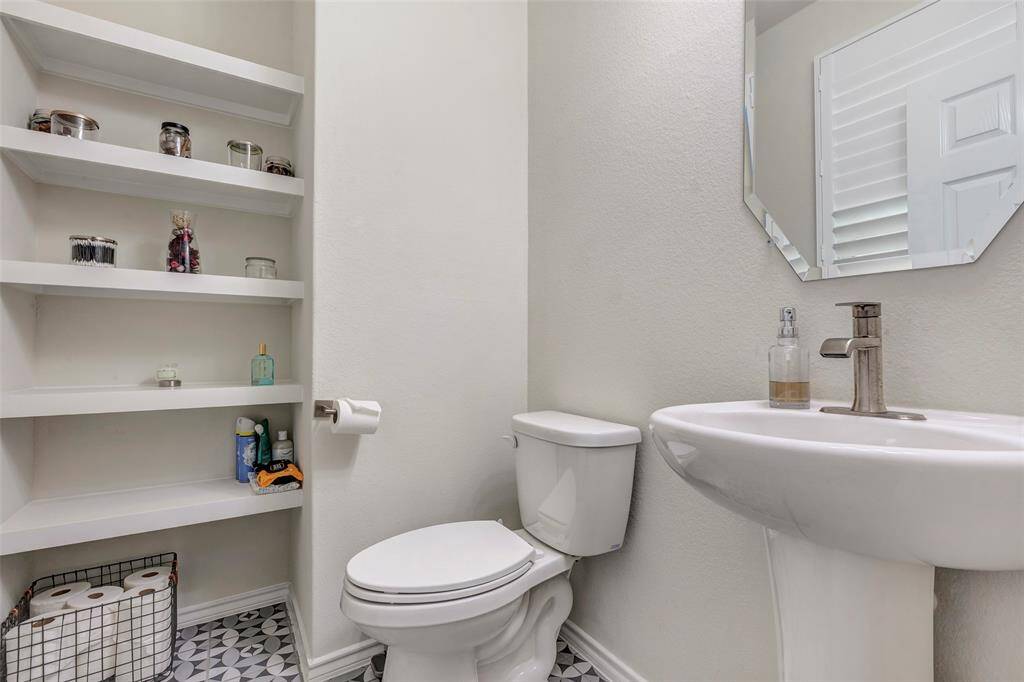 This is a compact, efficiently designed bathroom with built-in shelving for storage, a pedestal sink, and a toilet. The space features a neutral color scheme and patterned floor tiles, with a small window for natural light.