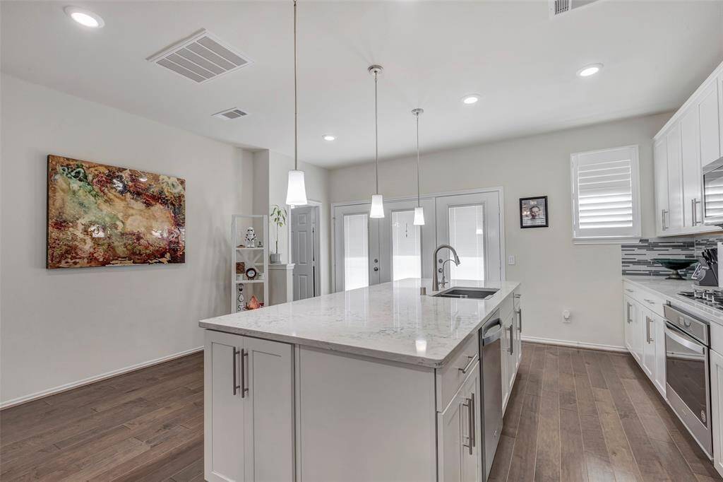 This is a modern, spacious kitchen with white cabinetry, stainless steel appliances, a large island with a sink, and pendant lighting. The room features hardwood flooring and recessed lighting, creating a bright and airy space.