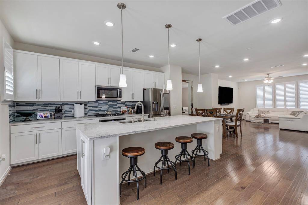 This is a modern, open-concept kitchen and living area featuring white cabinetry, stainless steel appliances, a large island with seating, and hardwood floors extending into a spacious living room with ample natural light. Landlord is supplying all appliances with lease agreement.