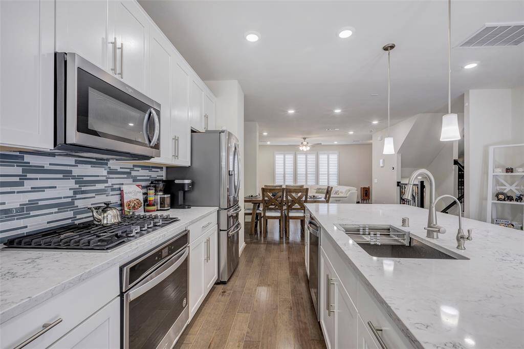 This is a modern, spacious kitchen with white cabinetry, stainless steel appliances, marble countertops, and a stylish backsplash. It features an island with a sink and a breakfast bar that opens up to a well-lit dining and living area.