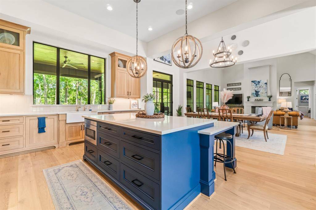 This photo highlights a spacious, modern open-concept kitchen and living area. A large island with bar seating, rich wooden cabinetry, and an abundance of natural light from expansive windows define the space. The adjoining dining and living areas are tastefully furnished, contributing to a warm and welcoming ambiance.