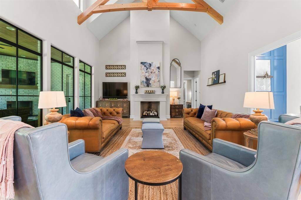 This spacious living room features high vaulted ceilings with exposed wooden beams, large windows for ample natural light, and a cozy fireplace.