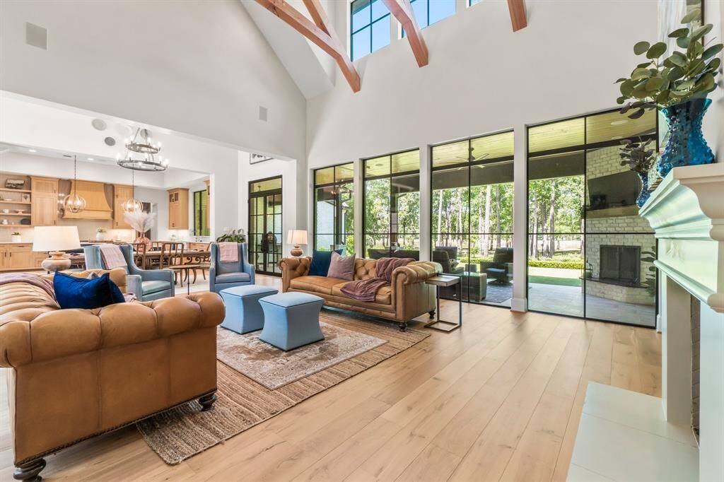 This expansive living room boasts soaring ceilings with exposed beams, oversized windows that flood the space with natural light, and an open layout seamlessly connecting to the kitchen and dining area. It also opens to a covered patio overlooking the serene, wooded backyard, blending indoor comfort with outdoor beauty.