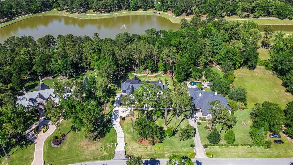 This aerial view showcases a spacious, elegant home nestled among lush greenery and mature trees. The property features a long driveway, beautifully landscaped gardens, and is situated near a serene lake with a scenic walking trail that winds around the water, offering a tranquil and private setting.