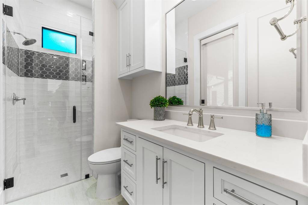 This modern bathroom features a sleek glass-enclosed shower with stylish tile accents, a white vanity with ample storage, and elegant fixtures. The bright space is complimented by a large mirror and contemporary finishes.