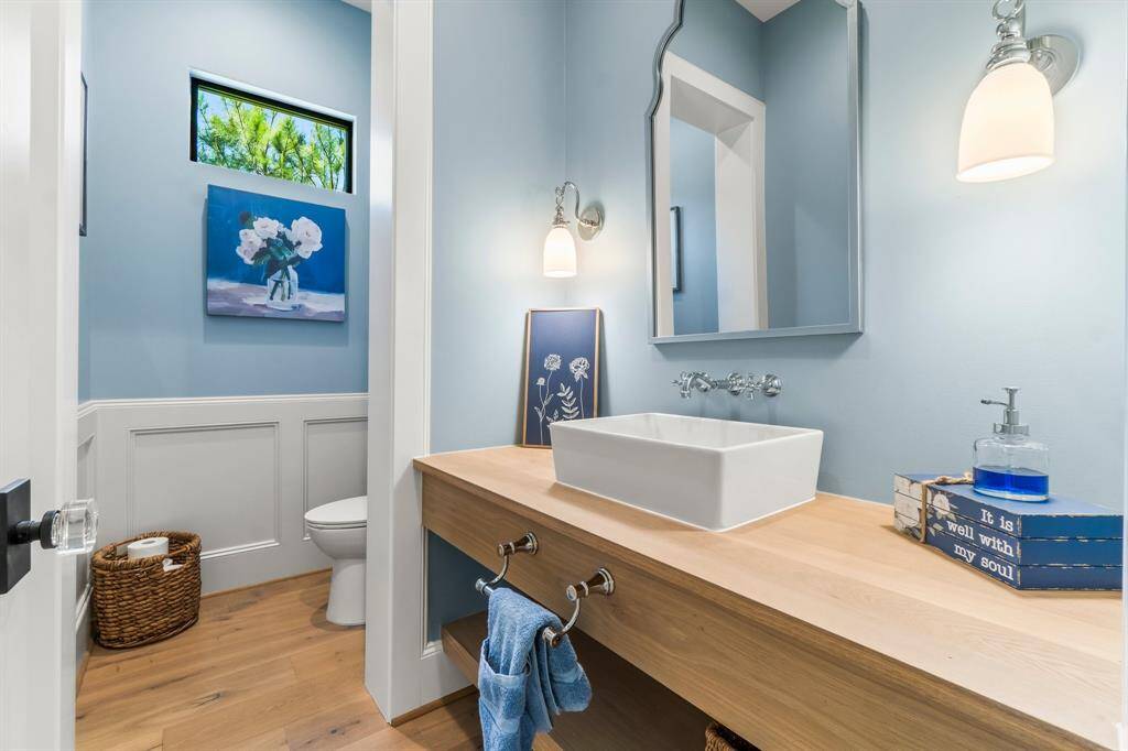 This photo showcases a bright and stylish bathroom with wainscoting. It features a modern sink with a wooden countertop, wall-mounted faucet, and elegant lighting fixtures.