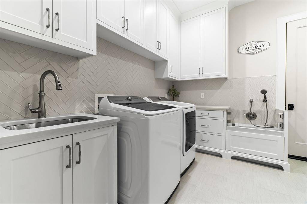 This is a modern, well-equipped laundry room featuring white cabinetry, a washer and dryer, a sink with a sleek faucet, and ample storage space. The design includes a herringbone tile backsplash and a pet washing station, combining functionality with style.
