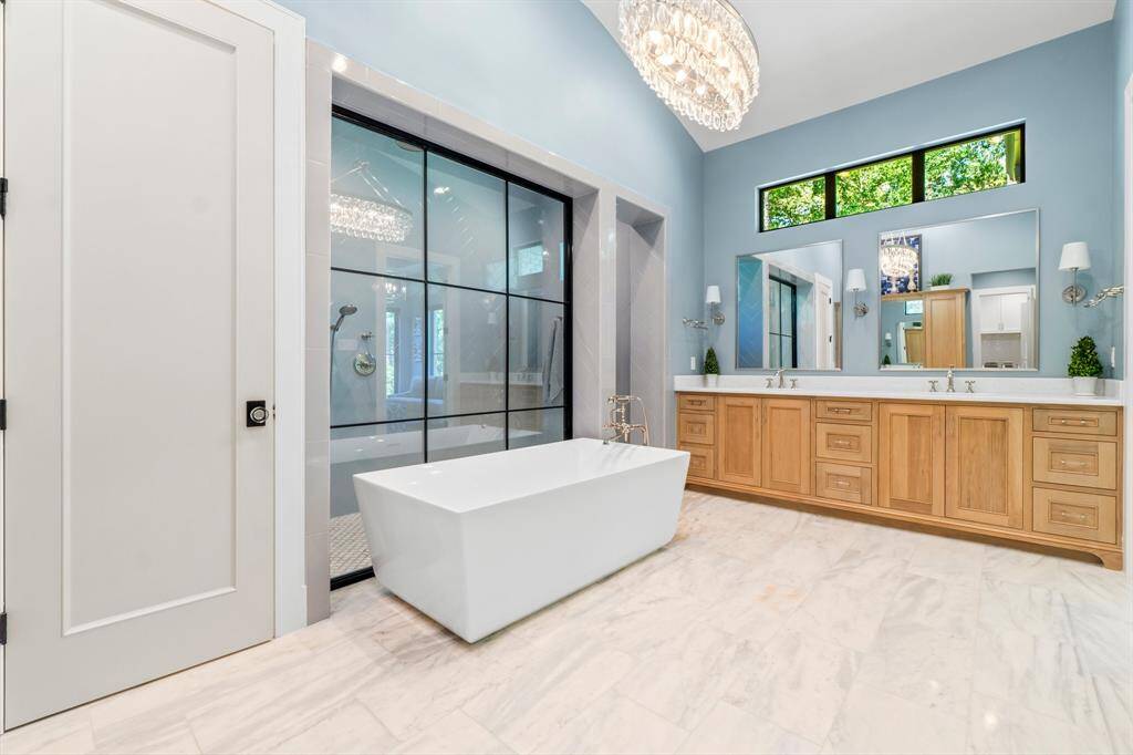This spacious bathroom features a modern freestanding tub, a large glass-enclosed shower, and a double vanity with ample storage. The room is brightened by a large window and elegant chandelier, providing a luxurious and inviting atmosphere.
