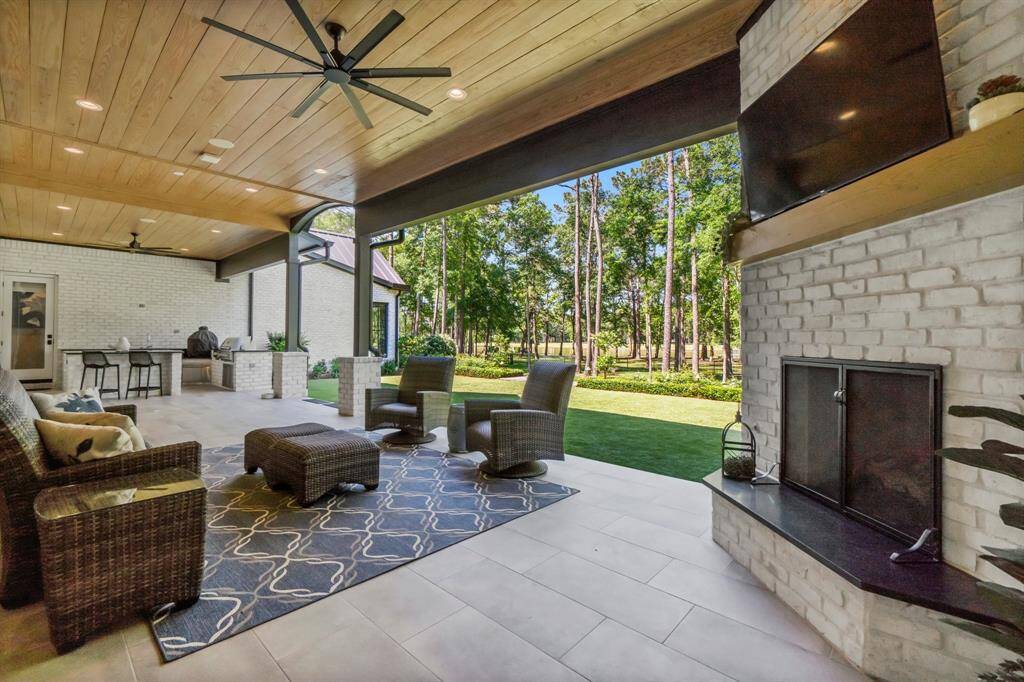 This spacious outdoor patio features a cozy seating area with a fireplace. The ceiling is beautifully paneled with wood, and large fans provide a breeze. Overlooking a lush, tree-filled yard, it's perfect for entertaining or relaxing.