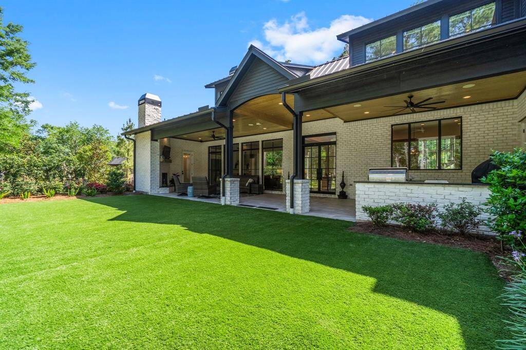 This photo showcases the spacious backyard with a covered patio featuring the built-in grill area and cozy seating area surrounds an outdoor fireplace. The well-maintained lawn and lush surrounding greenery create a private, serene space perfect for both relaxation and entertaining.