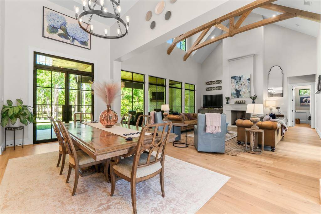 This spacious living and dining area features high vaulted ceilings with exposed wooden beams, large windows offering abundant natural light, and elegant hardwood floors. The open layout includes a cozy seating area with a fireplace, perfect for entertaining or relaxing.