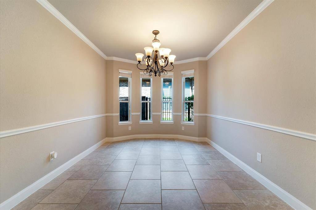 This inviting dining room features neutral walls with elegant crown molding and a classic chandelier. The windows provide ample natural light, and the tiled floor adds a modern touch, making it perfect for a dining area or formal sitting room.
