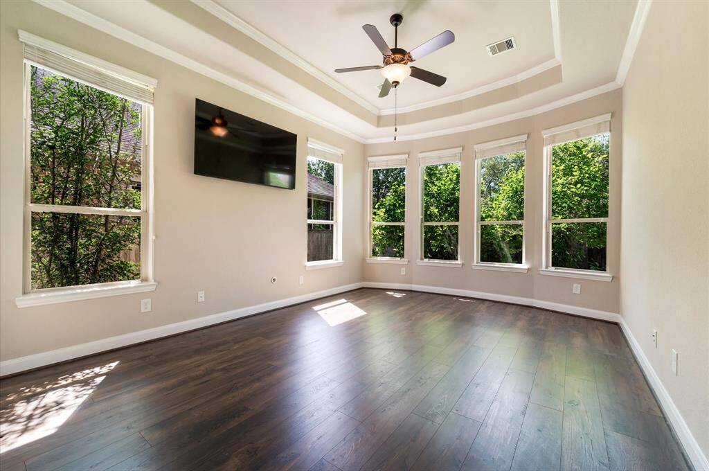 Bright and spacious Primary Bedroom with large windows, tray ceiling and crown molding offering plenty of natural light. It features beautiful LVP floor for easy low maintenance, a ceiling fan, and a mounted TV, creating a warm, airy space and perfect ambience to relax.