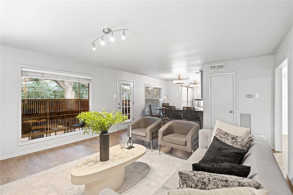 The living area overlooks a private balcony through large picture windows. The door resting between the living room and kitchen provides its access.