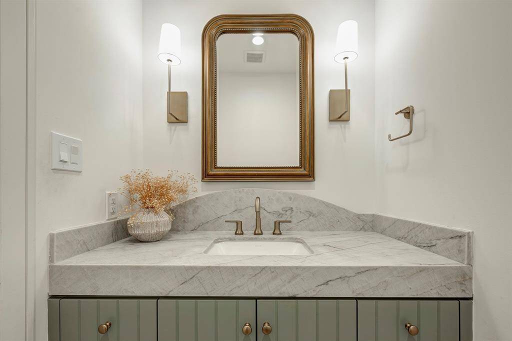 Elegant powder room features custom v-groove cabinetry with a soft, leathered quartzite countertop. Champagne bronze faucet and matching hardware add warmth and refinement, while dual wall sconces create flattering ambient light.