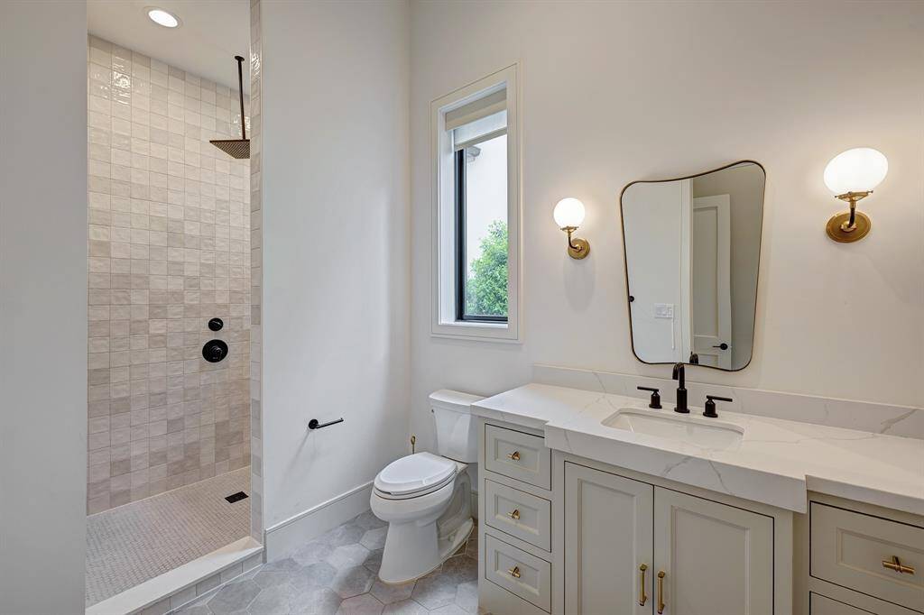 Downstairs private guest bath features a large walk-in tile shower with a rain showerhead in a sleek matte black finish. A Statuario quartz countertop is paired with a designer vanity mirror and two elegant wall sconces, while matte-finish hexagon tile flooring adds a stylish and modern touch.