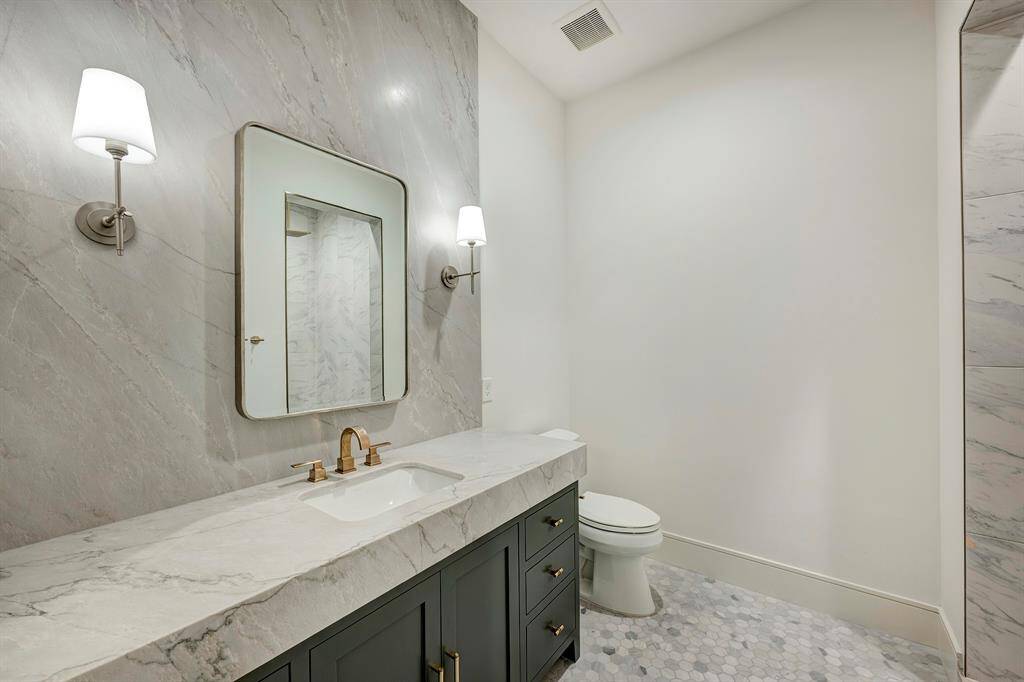 Cool and sophisticated ensuite bath (also accessible from the gameroom) features counter-to-ceiling elegance with a leathered white Santorini quartzite countertop and full-height backsplash. Custom inset cabinetry provides ample storage, complemented by a spacious walk-in shower with porcelain surround and built-in shampoo niche. Classic marble hexagon tile flooring anchors the space, while brushed nickel wall sconces and a designer mirror complete the refined look.