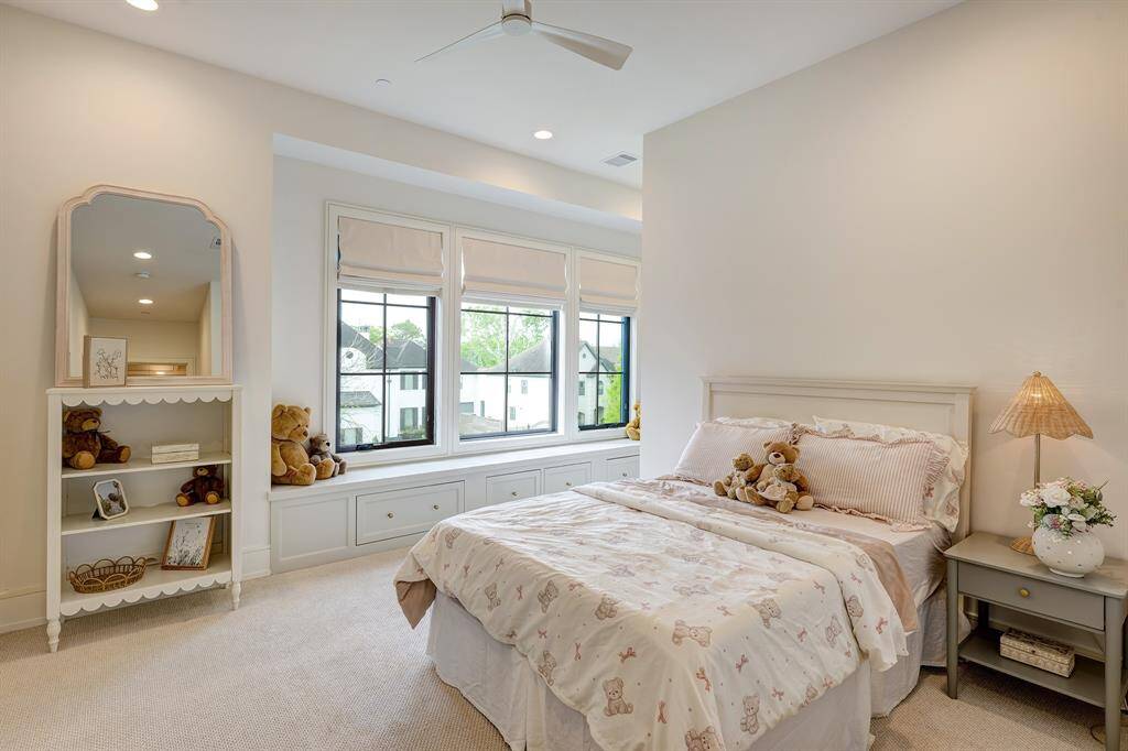 This spacious (and super cute) secondary bedroom is filled with natural light from a trio of large windows overlooking the front lawn. Thoughtfully designed with comfort and functionality in mind, it features plush carpeting, a built-in window seat with storage drawers, and custom Roman shades from The Shade Store. A ceiling fan provides added comfort, and the layout easily accommodates both rest and play.