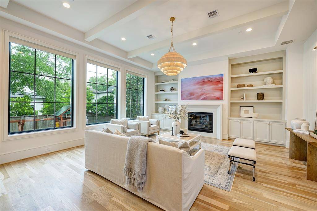 This light-filled sanctuary blends elegance with comfort. The living room is centered around a dramatic cast-stone gas fireplace, flanked by built-in cabinetry and flooded with natural light from four oversized picture windows overlooking the backyard. The painted ceiling beams and tongue-and-groove art niche contribute architectural detail, complemented by practical floor outlets and sophisticated lighting.
