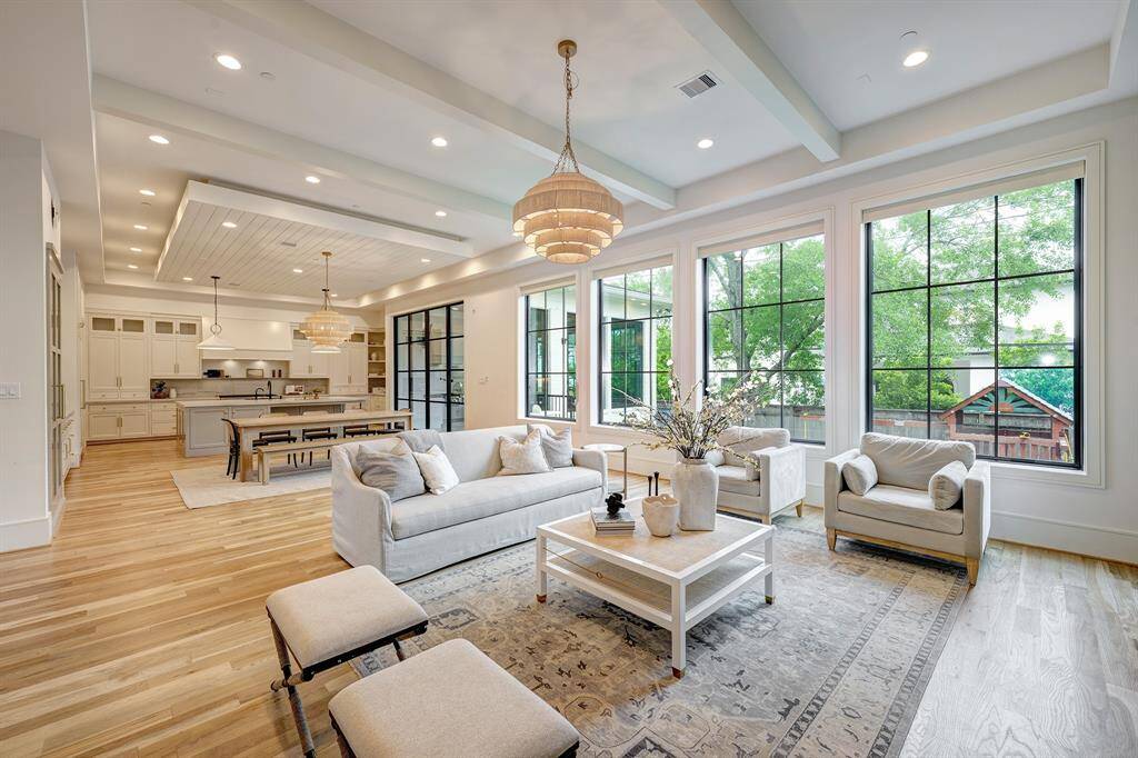 The expansive open-concept living, dining, and kitchen area creates an ideal space for both intimate evenings and grand gatherings. Four large Andersen windows merge indoor and outdoor spaces seamlessly while offering tranquil backyard views. The elegant atmosphere flows from soft neutral tones, white oak floors, and a painted beam ceiling, while a striking tiered chandelier adds a dramatic touch of glamour.