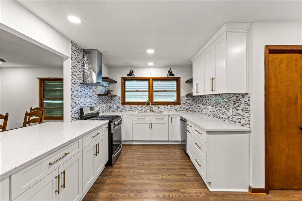 This kitchen features sleek white cabinetry, a stylish mosaic tile backsplash, and stainless steel appliances. It offers ample counter space with a large peninsula and dining area nearby.