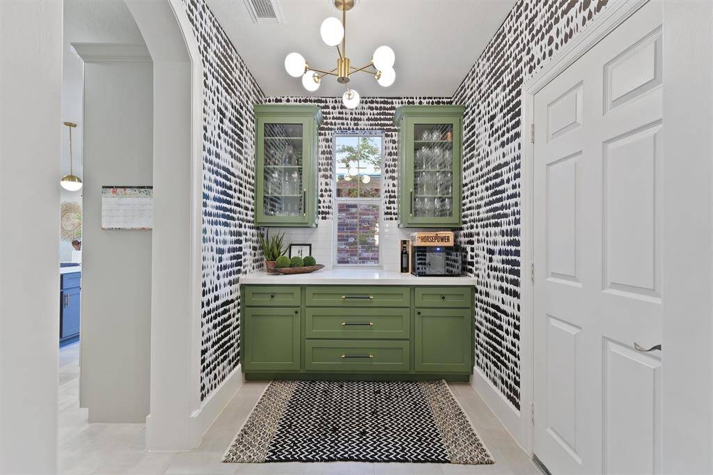 The custom dry bar features stunning green cabinetry, quartz countertops, and glass paned cabinet doors. The modern light fixture, bold wallpaper, and window view makes this area a fun, flexible space beyond the kitchen.