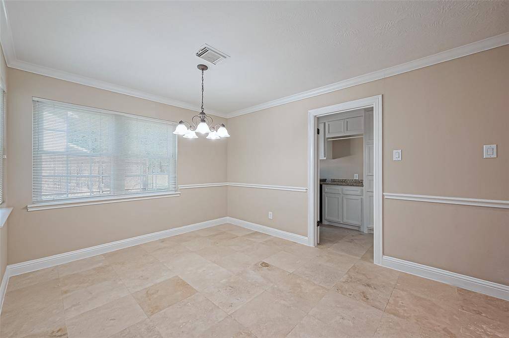 This bright dining area features neutral tones, large windows with blinds, Travertine flooring, and a classic chandelier. It opens into a kitchen with access to the laundry room and 1/2 bath.