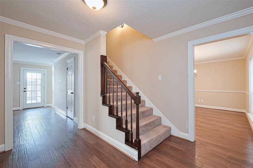 This inviting entryway features warm engineered wood flooring and neutral walls. It includes a carpeted staircase with dark wood railings leading upstairs. The space is well-lit with natural light from adjacent rooms, including a room with French doors and another with a chandelier, ideal for dining or a home office.