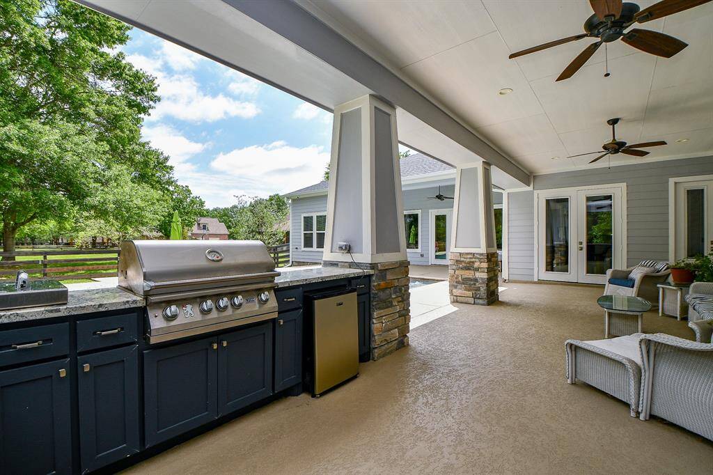 The built-in outdoor kitchen featuring a sink, SS grill and mini fridge. The area includes comfortable seating space, perfect for entertaining, with a view of a lush backyard and the sparkling pool.