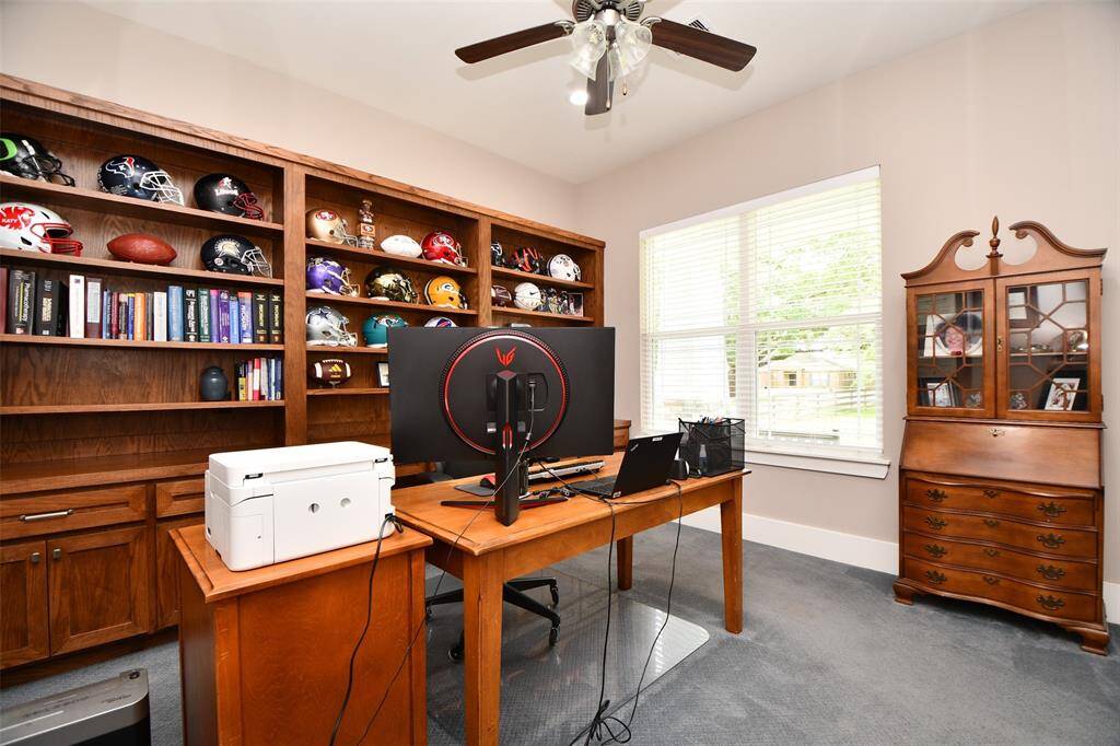 The study/home office features a full wall of built-in wood shelves, a large window for natural light, and ceiling fan creating a cozy and functional workspace.