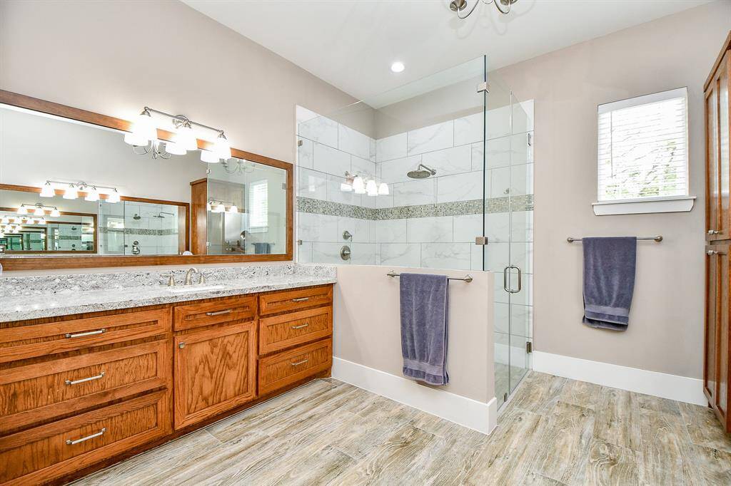 This view of the primary bathroom showcases the large glass-enclosed shower and double vanity with granite countertops. Warm wood-look flooring and bright modern lighting create a spa-like, inviting feel.