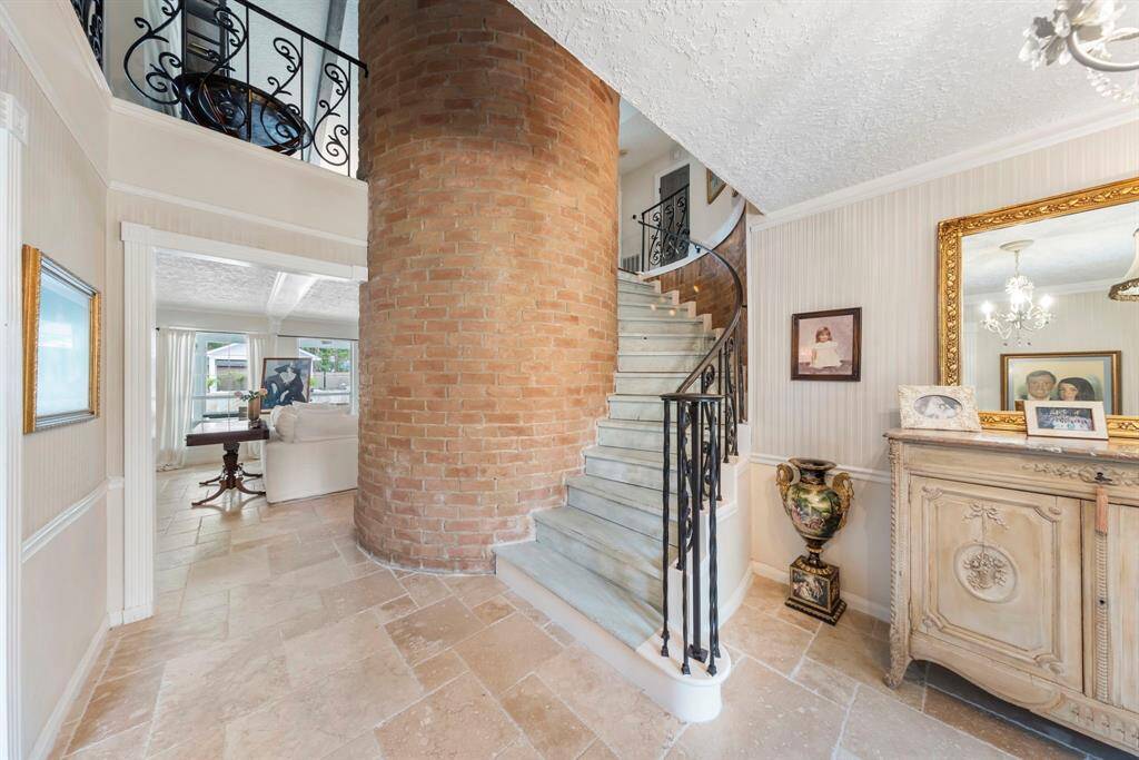 Step inside to timeless traditional charm featuring elegant tile flooring, intricate crown molding, and a striking iron spindle staircase that frames the dramatic two-story brick fireplace.