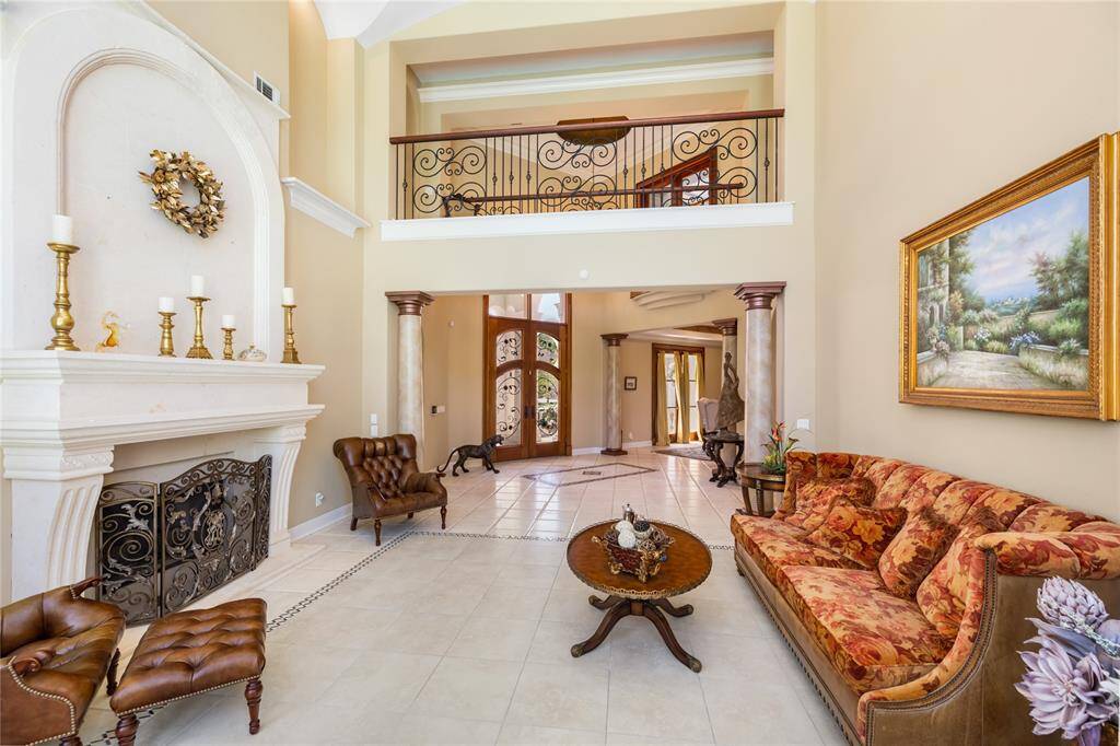 This elegant living room features high ceilings and a grand fireplace. A wrought iron balcony overlooks the space, complemented by large windows that allow natural light to fill the room.