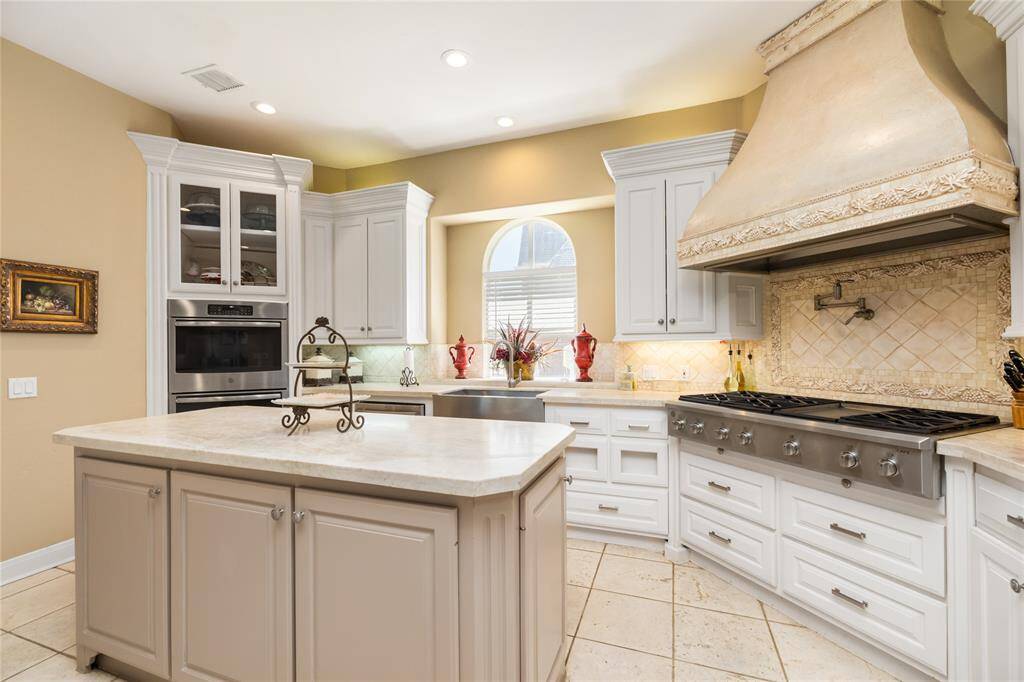 The kitchen boasts a generous layout with stylish cabinetry, a large island, and top-of-the-line double ovens. The six-burner stove, complemented by an ornate range hood, is a striking focal point. Natural light pours in through the arched window, accentuating the intricate tile backsplash and the serene, neutral tones that flow throughout the space.