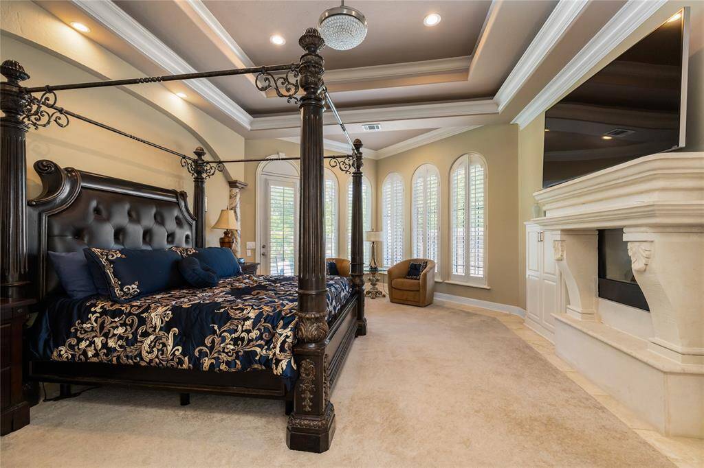 The luxurious primary bedroom is adorned with elegant tray ceilings, creating a serene retreat. A cozy seating area by the tall, arched windows invites relaxation, while the stylish fireplace adds warmth and charm, enhanced by abundant natural light.