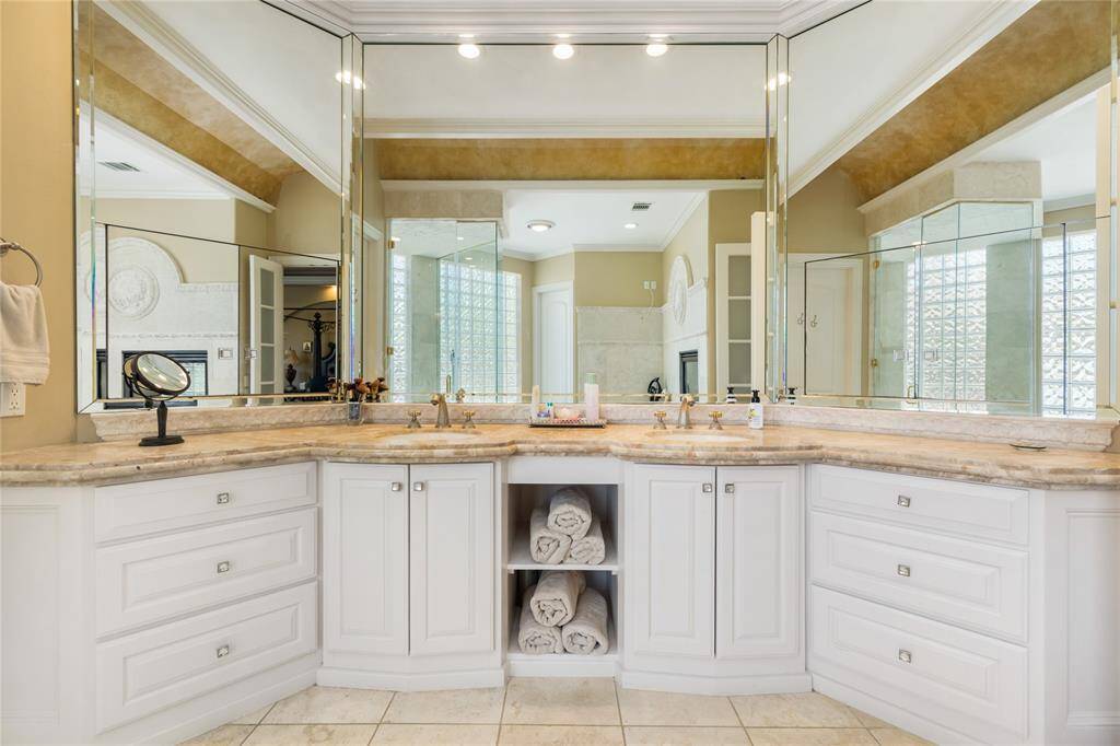 This bathroom features a spacious double vanity with elegant white cabinetry and marble countertops. Large mirrors enhance the space, and ample storage is available with drawers and shelves. The neutral color palette and high-end fixtures create a luxurious and inviting atmosphere.
