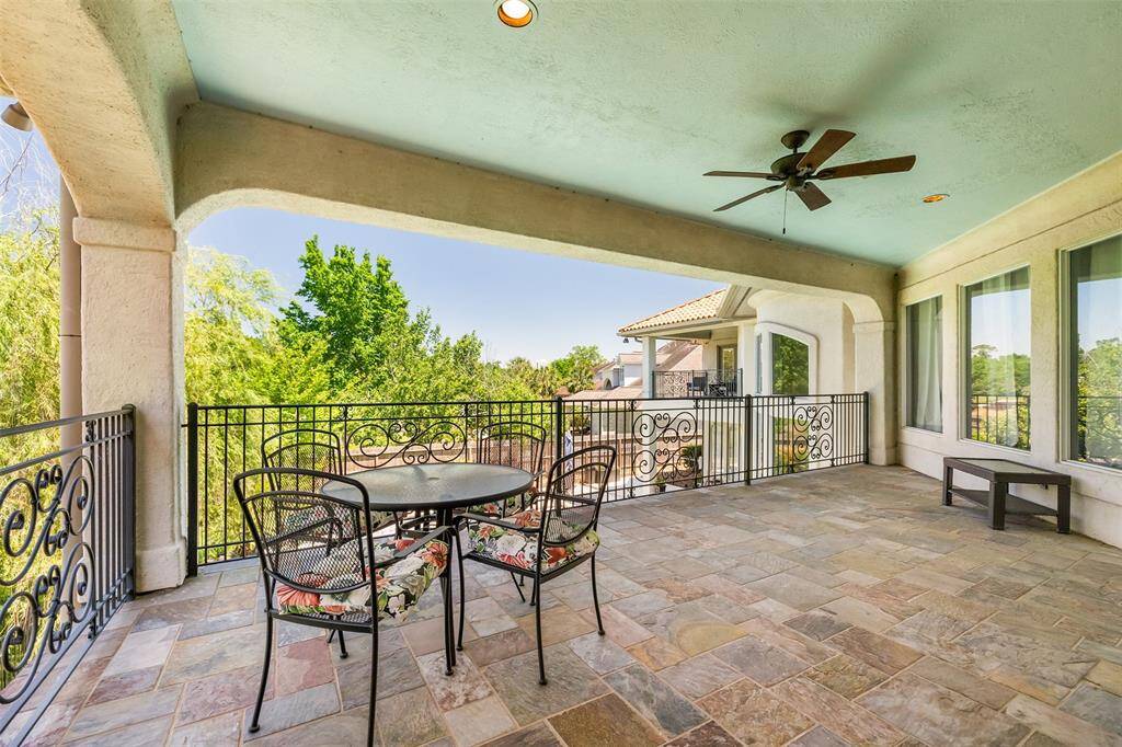 This spacious covered balcony features elegant wrought iron railings, a ceiling fan, and a tiled floor. It offers a serene view of lush green trees, perfect for outdoor dining or relaxation.