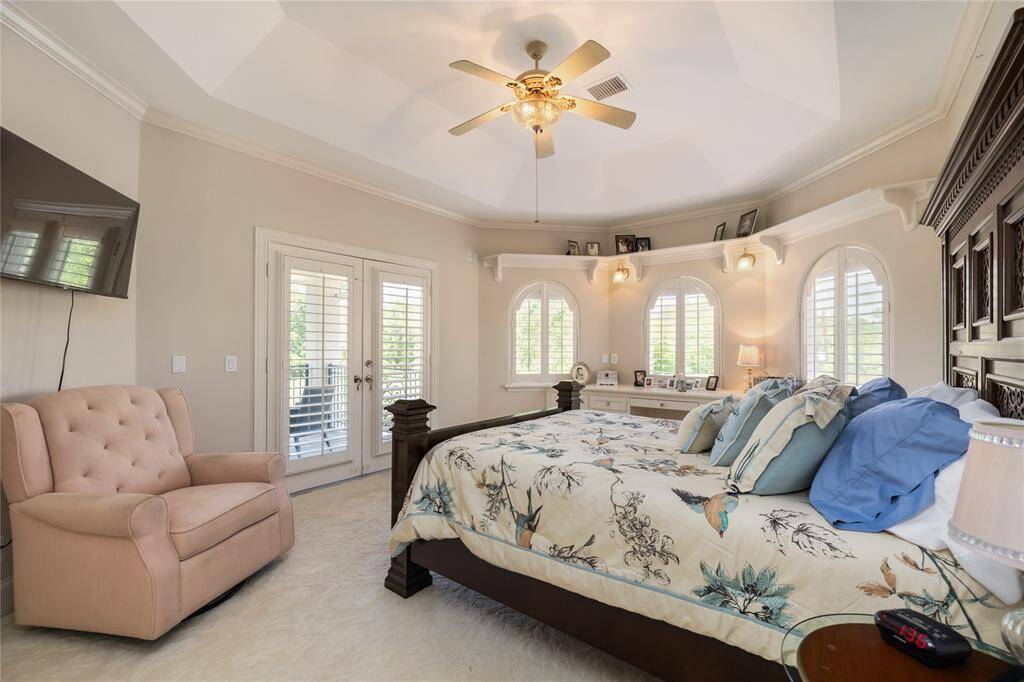 This spacious bedroom features a vaulted ceiling with a ceiling fan, large windows with plantation shutters, and French doors leading to a balcony. It includes a cozy seating area and built-in shelves, creating a bright and inviting atmosphere.