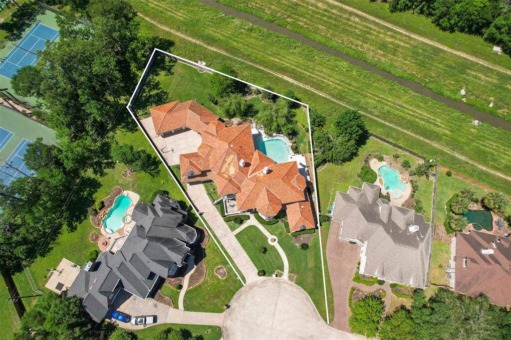 This aerial view highlights the luxurious layout of the property, featuring a terracotta roof and expansive outdoor space, including a large pool. Surrounded by lush greenery, the home is situated in a vibrant neighborhood with nearby tennis courts.