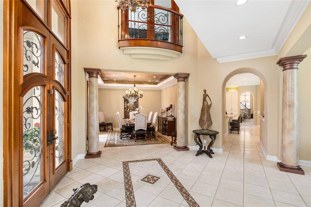 This elegant foyer features a grand entryway with impressive double doors featuring decorative ironwork. The space opens into a formal dining area with columns and an ornate tray ceiling, while a curved staircase leads to the upper level. The design is classic and sophisticated, with tiled flooring and warm tones throughout.