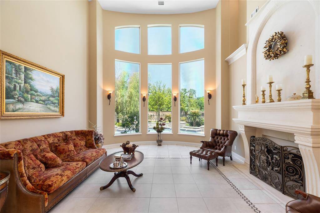 This elegant living room features soaring ceilings and large arched windows, offering abundant natural light and a view of the landscaped garden and pool. It includes a cozy fireplace, creating an inviting and luxurious atmosphere.
