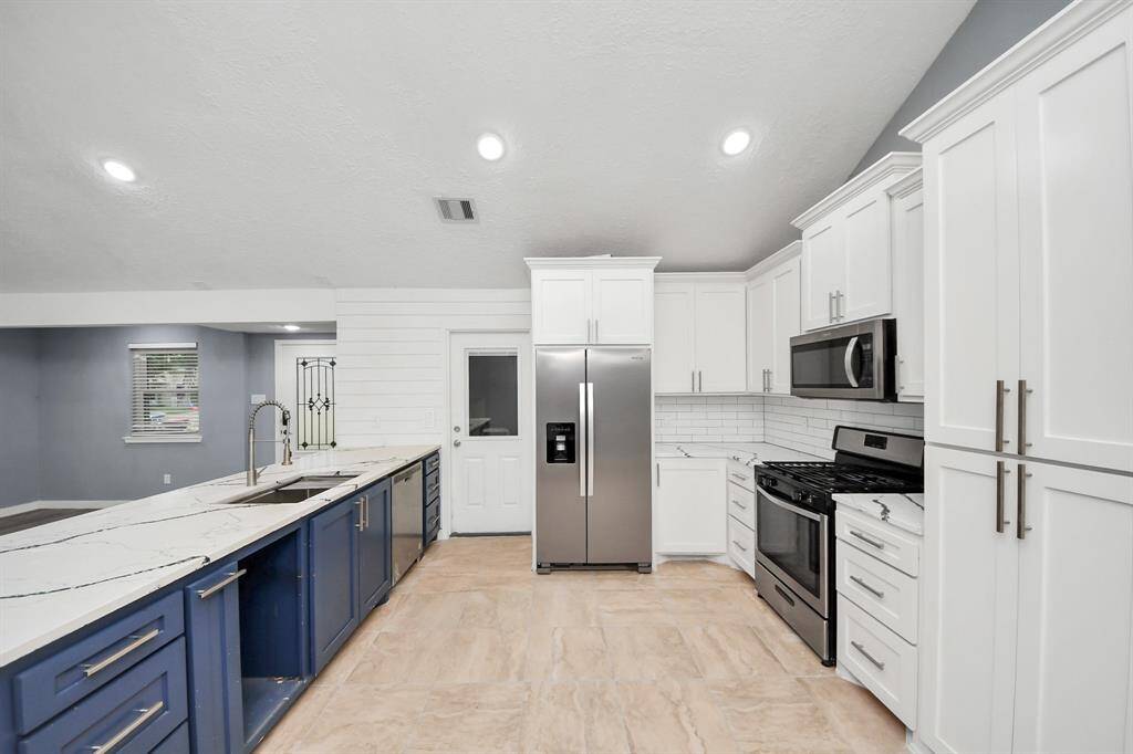 This modern kitchen features sleek white cabinetry, a stainless steel refrigerator and stove, a spacious marble countertop with a deep sink, and a stylish blue island. The neutral palette and recessed lighting create an inviting, airy atmosphere.