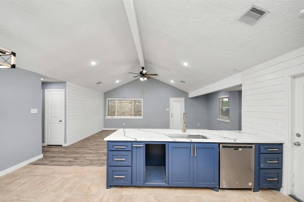 This modern kitchen features a spacious open layout with vaulted ceilings, a central island with blue cabinetry, and stylish marble countertops. The room is well-lit with recessed lighting and offers a ceiling fan for comfort. Light gray walls and wood-look flooring add a contemporary touch.