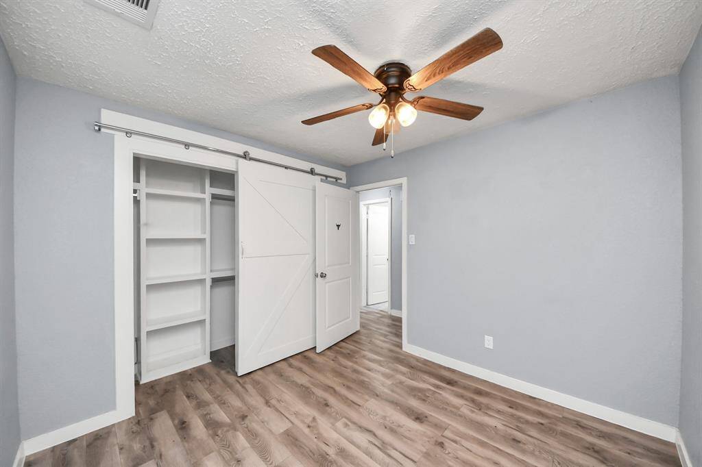 This room features a modern design with light gray walls, a ceiling fan with wooden blades, and wood-look flooring. It includes a sliding barn door leading to a closet with built-in shelving, offering ample storage space. Ideal for a cozy bedroom or home office.