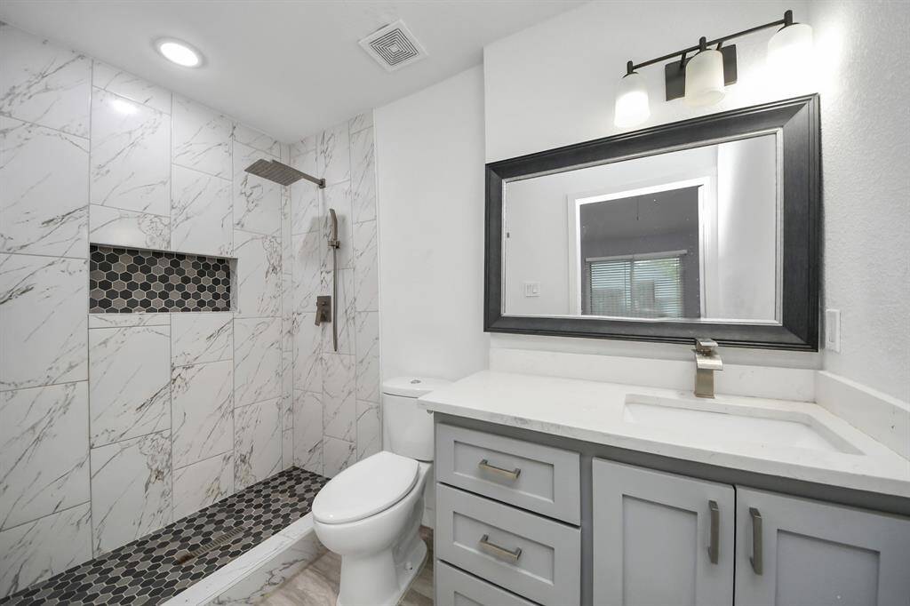 Modern bathroom featuring a spacious walk-in shower with elegant marble-style tiles and a contrasting hexagonal tile accent. The vanity includes a large mirror, sleek sink, and ample storage with gray cabinets, all illuminated by stylish lighting.
