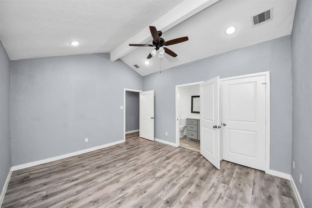 This room features a vaulted ceiling with a modern ceiling fan, laminate wood flooring, and soft gray walls. It has an en-suite bathroom with a visible vanity and is well-lit with recessed lighting. The space feels open and inviting.