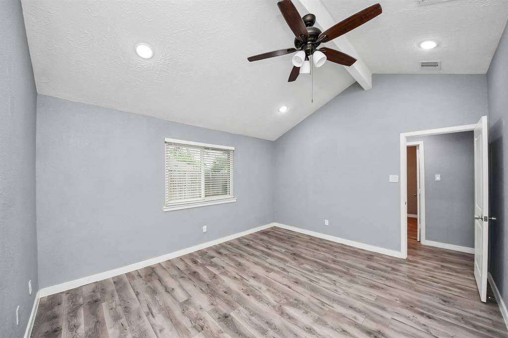 This room features a vaulted ceiling with a central fan, soft gray walls, and wood-look flooring. It has a window for natural light and an open doorway leading to another area, creating a spacious and inviting atmosphere.