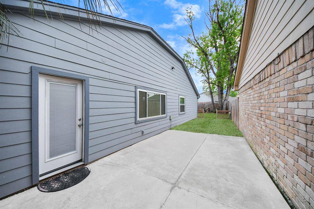 This photo shows a tidy backyard area with a concrete patio leading to a grassy section. The house exterior is gray siding with a brick wall on one side, and a door opens into the patio. It's a compact space ideal for outdoor seating or a small garden.