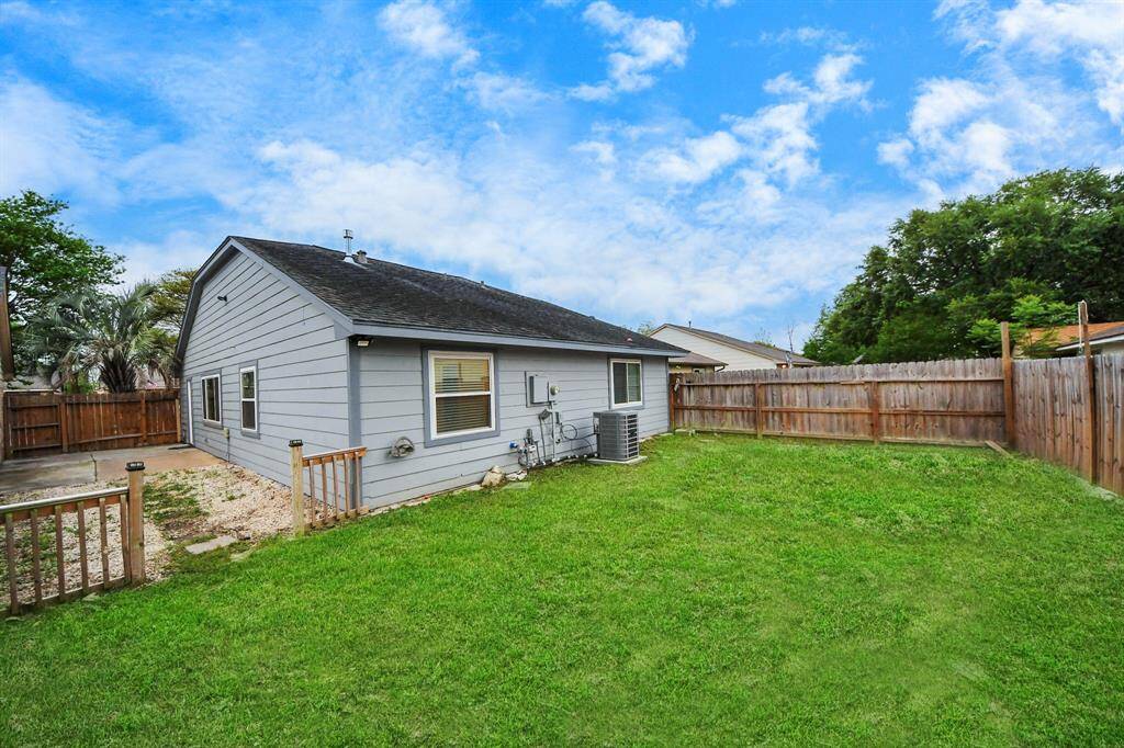 This photo shows a cozy, single-story home with grey siding and a fenced backyard. The yard features a grassy area and a small paved section, ideal for outdoor activities. The setting is private, with wooden fencing and mature trees in the background.